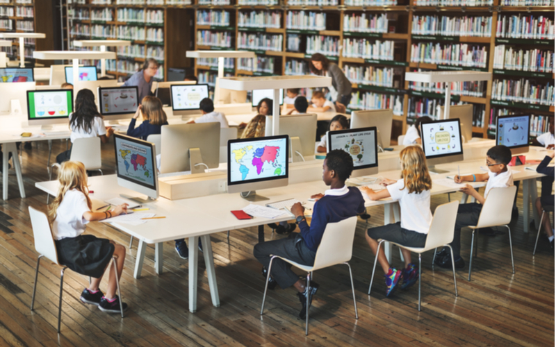 Classroom of students sat in a library around desks with each at their own desktop computer