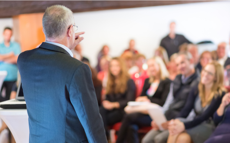 Behind shot of a man in a suit addressing a room of people