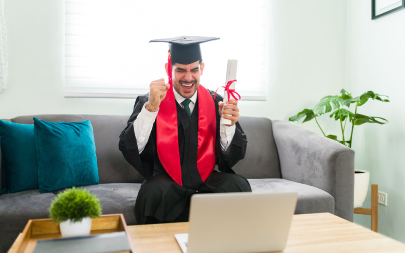 Latin young man feeling very excited to receive his bachelor's degree. Male graduate in a gown shouting with excitement while sitting at home during a virtual graduation